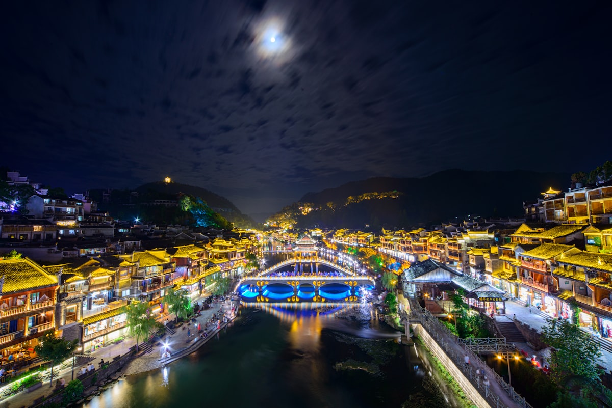 Fenghuang old town glowing at night along the Tuo River beneath a moonlit sky