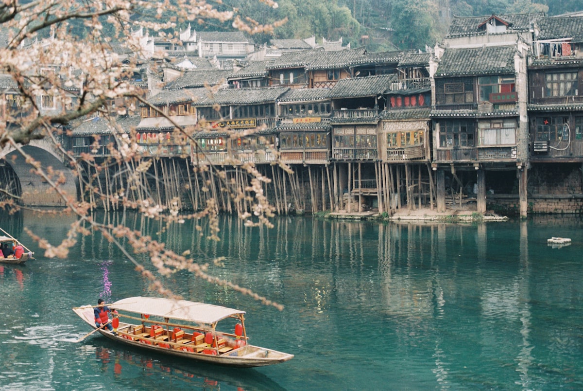 A covered tour boat glides past wooden stilt houses on the Tuo River in Fenghuang