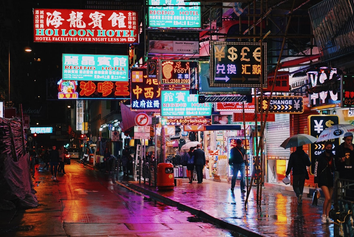 Neon signs glow over a rainy Kowloon street as pedestrians pass with umbrellas