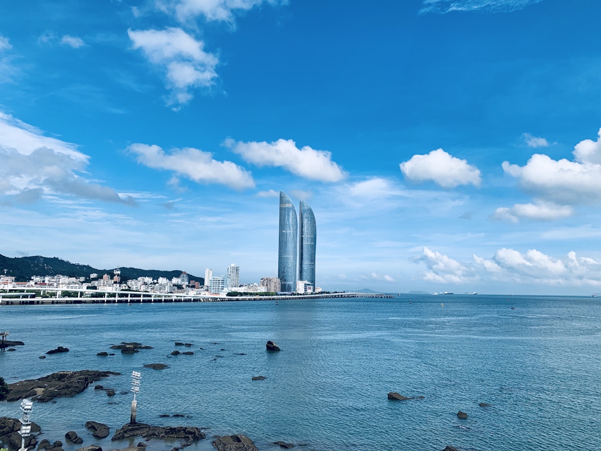 Twin Shimao Straits Towers rise over Xiamen's coastline, seen from a rocky beach