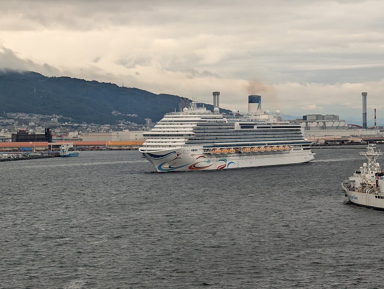 The Adora Magic City alongside at the port of Kobe, Japan, its rows of balcony cabins stacked above the hull.