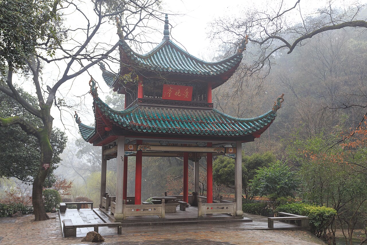 A two-tiered pavilion with green glazed roofs and red pillars, a gold-lettered red plaque beneath the upper eaves, stone benches around a stone table inside, and bare winter branches on a misty wooded slope behind