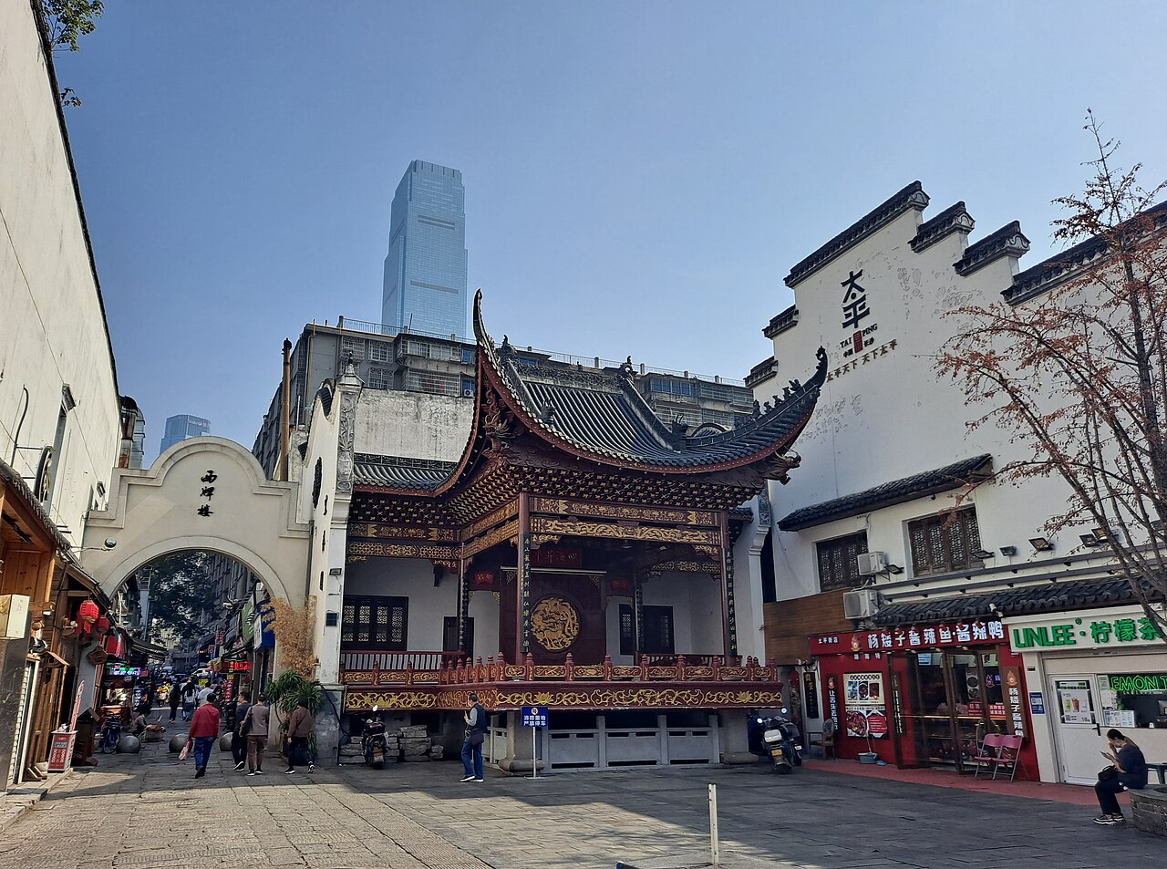 An ornate timber opera stage with an upswept black tiled roof and gilded carving stands in an open square, a whitewashed gable lettered in Chinese above the shopfronts to the right, a stone archway leading away to the left, and a glass tower rising beyond the rooftops