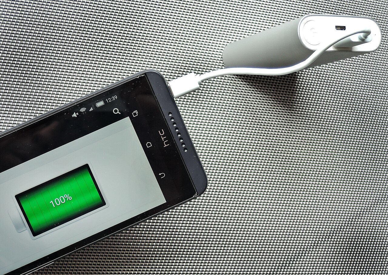 A smartphone connected to a slim silver USB power bank, charging on a wooden desk.
