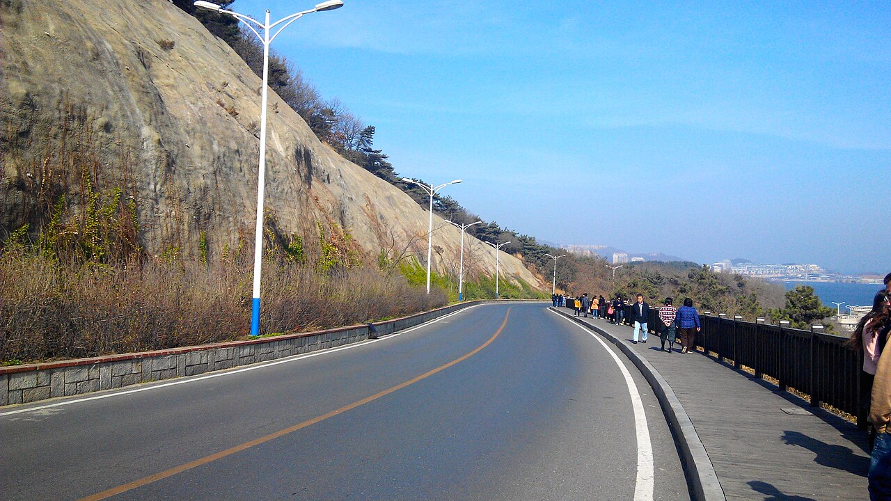 Binhai Road curving along a grassy cliff above the sea, a few walkers on the shoulder.