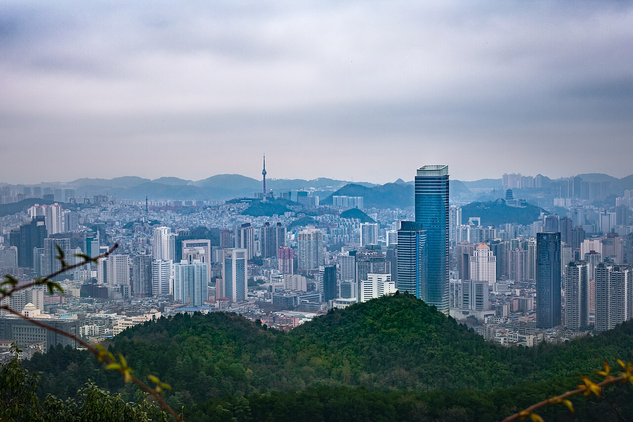 The Guiyang skyline seen from nearby mountains, with high-rise towers rising above the green hills of Guizhou.