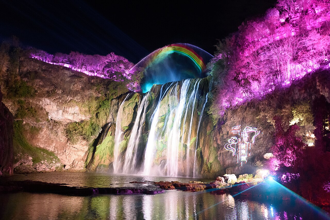 Huangguoshu waterfall illuminated in colour at night, with the cascade lit purple and gold above the river.