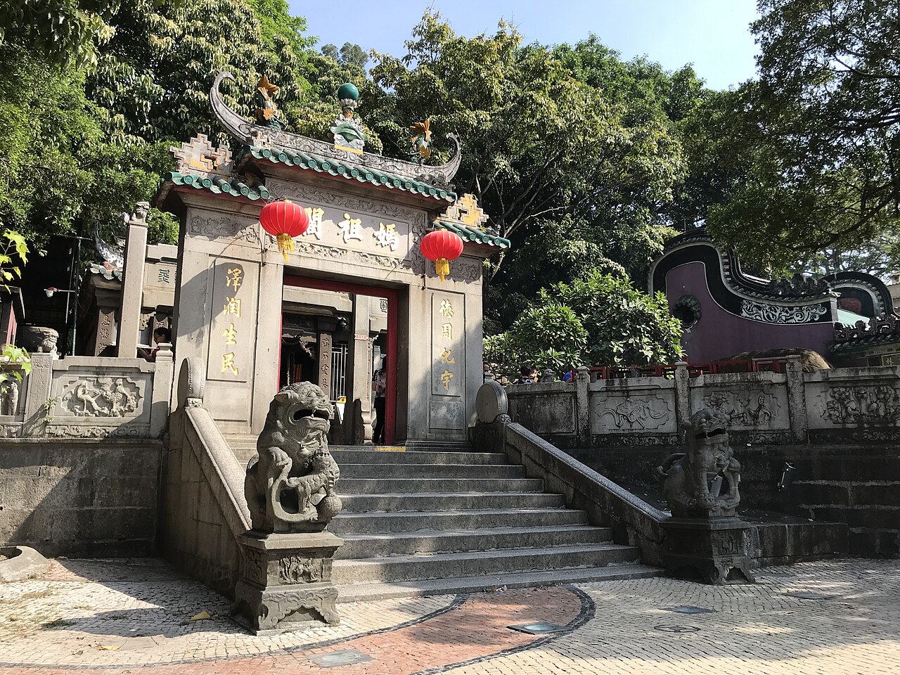 The stone gateway of A-Ma Temple under a green-tiled roof, hung with two red lanterns and flanked by carved stone lions at the top of the steps.