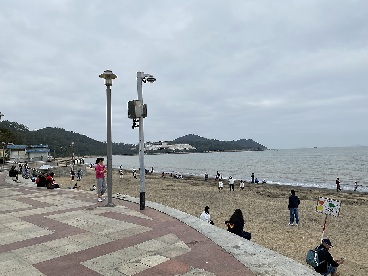 A wide stretch of dark sand under an overcast sky, with a tiled promenade in front and green headlands across the bay.