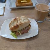 A pork chop in a crusty split roll on a white plate, with a glass of milk tea and a slice of honeyed toast behind it.