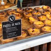 Trays of egg tarts behind bakery glass, their custard tops blistered and caramelised, beside a card reading Pastel de Nata.