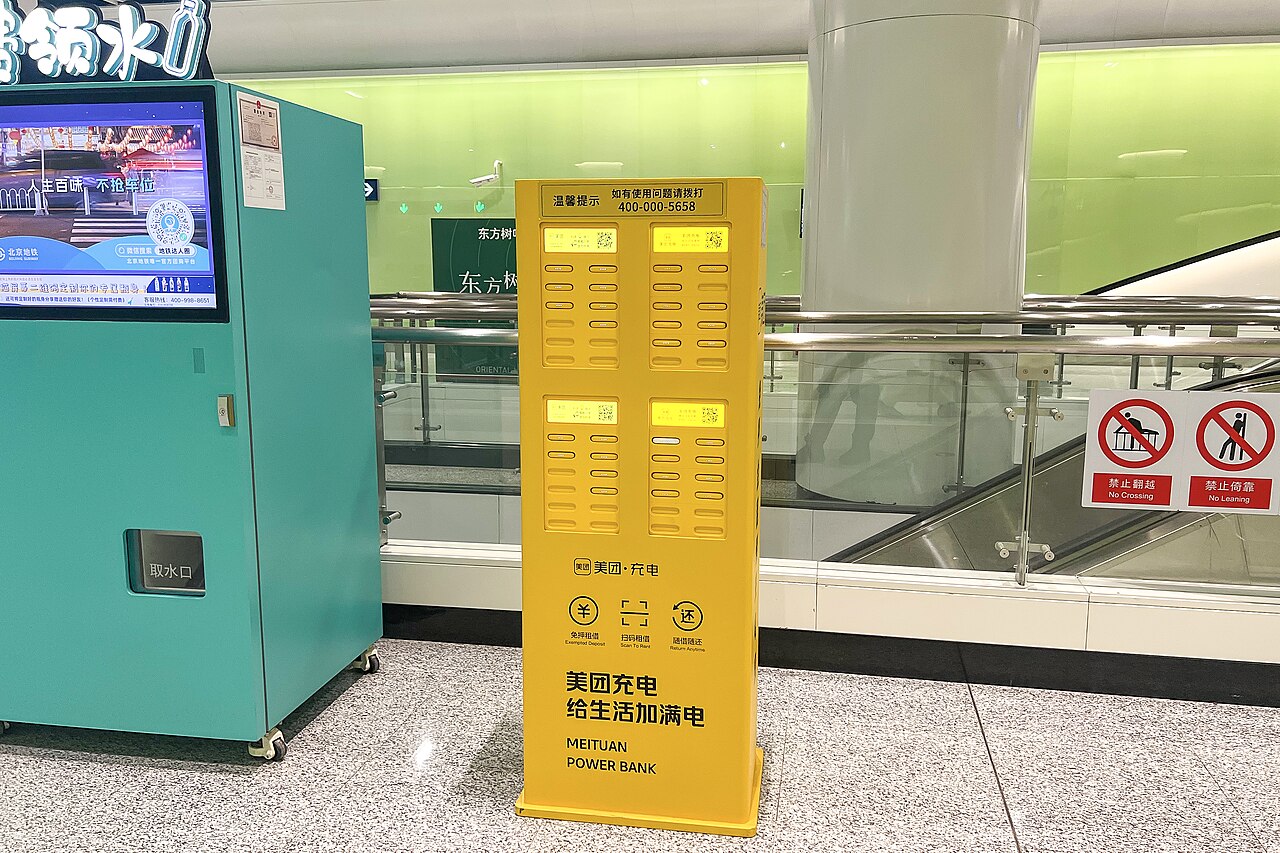 A yellow Meituan shared power bank rental kiosk on the concourse of a Beijing metro station, with rows of power bank slots and a QR code panel.