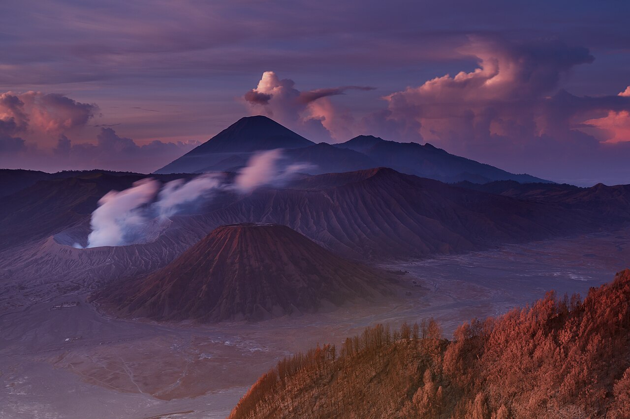 Mount Bromo volcano at sunrise, its smoking crater rising from a sea of sand with ridged volcanic cones around it.