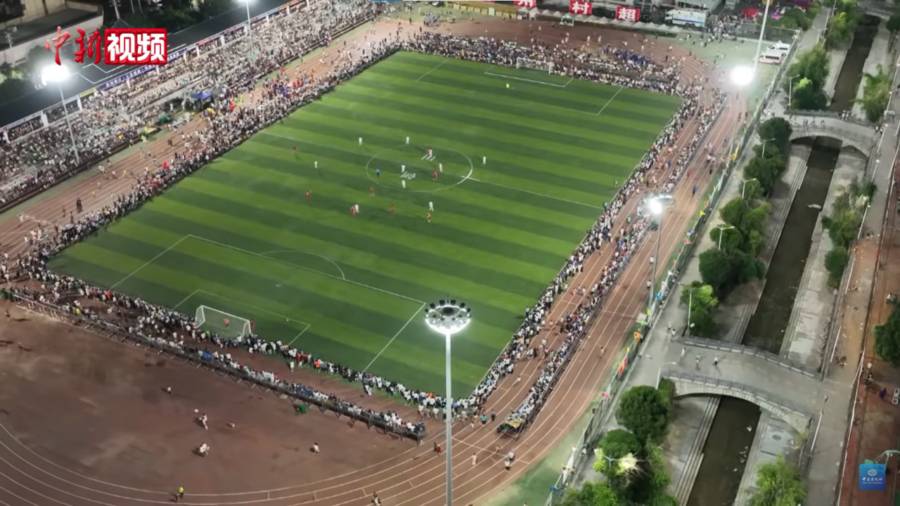 A packed village football stadium in Rongjiang, Guizhou, with crowds filling the stands around a small pitch during a Village Super League match.