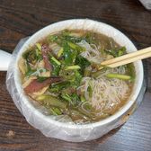 Thin rice noodles under a thick brown gravy with greens and slices of cured meat, in a plastic-lined bowl with chopsticks laid across.