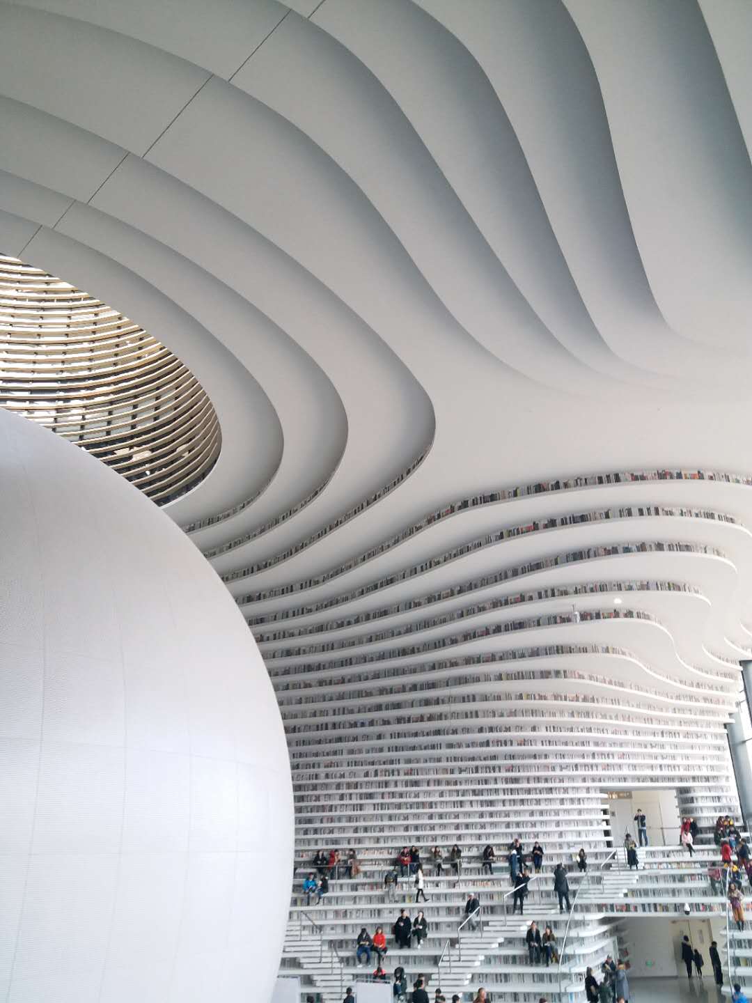 The white terraced book-mountain inside Binhai Library, readers scattered across its curved steps around a central sphere.