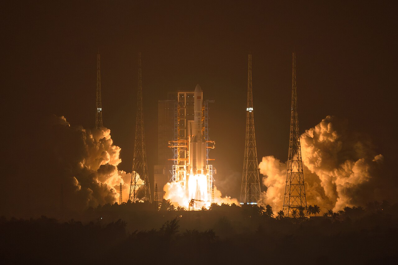 A Long March 5 rocket lifting off at night from Wenchang, the pad lit orange by the exhaust, with four lightning towers and a treeline of palms around it.