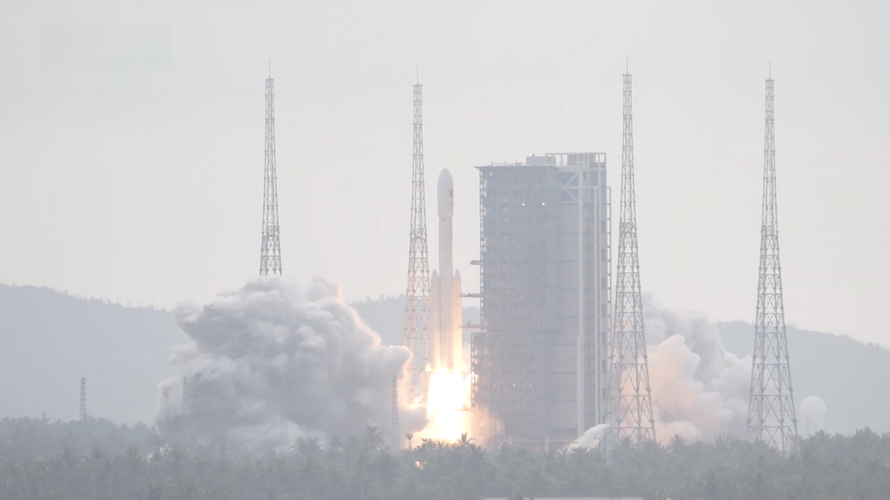 A Long March 7 rocket rising past its grey service tower in daylight, with wooded hills behind and coconut palms across the foreground.