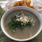 A bowl of thick grey fish broth topped with spring onion and tiny dried shrimp, with fried dough sticks in a bag behind it.