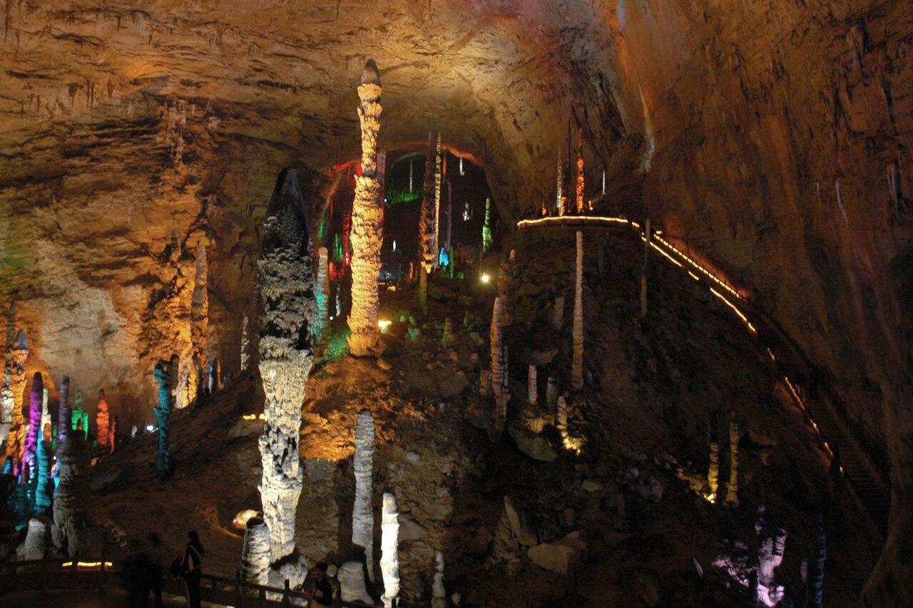 Colourful flood-lit stalactites rising from the floor of a vast limestone cave, with a walkway tracing an underground pool.