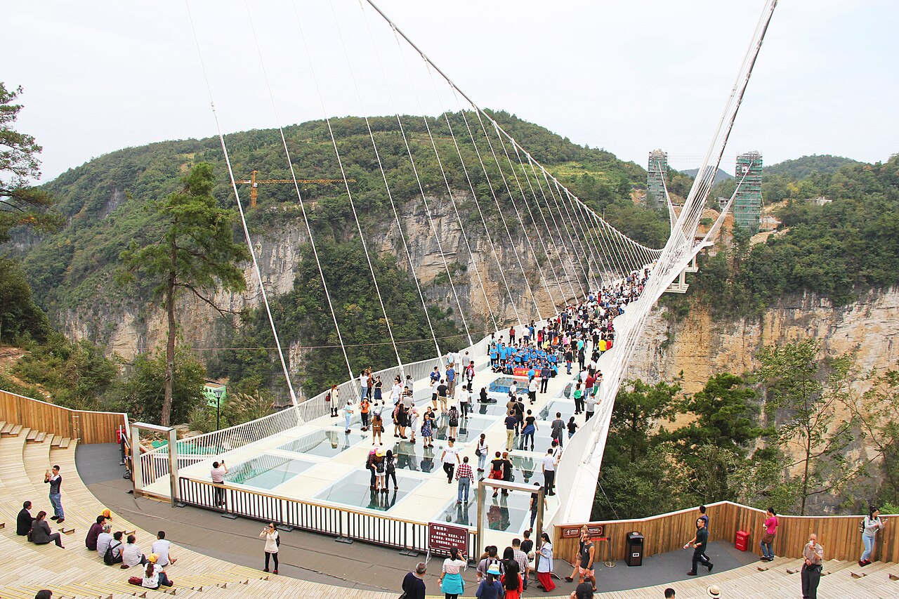 Crowded walkway on a suspension bridge with a transparent glass deck strung between sandstone cliffs above a forested gorge.
