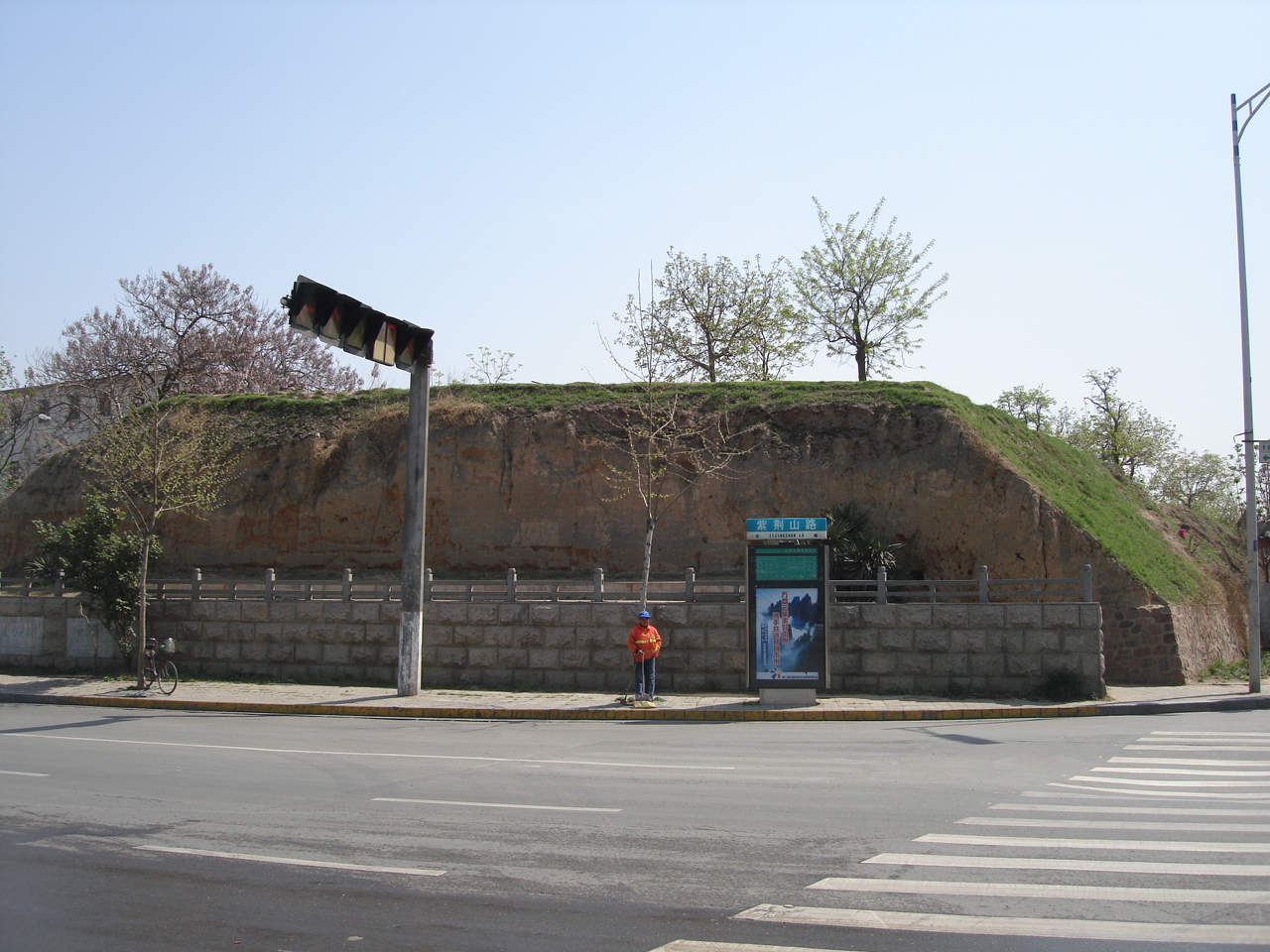 A grass-covered earthen Shang dynasty wall crossing a modern street corner in Zhengzhou.