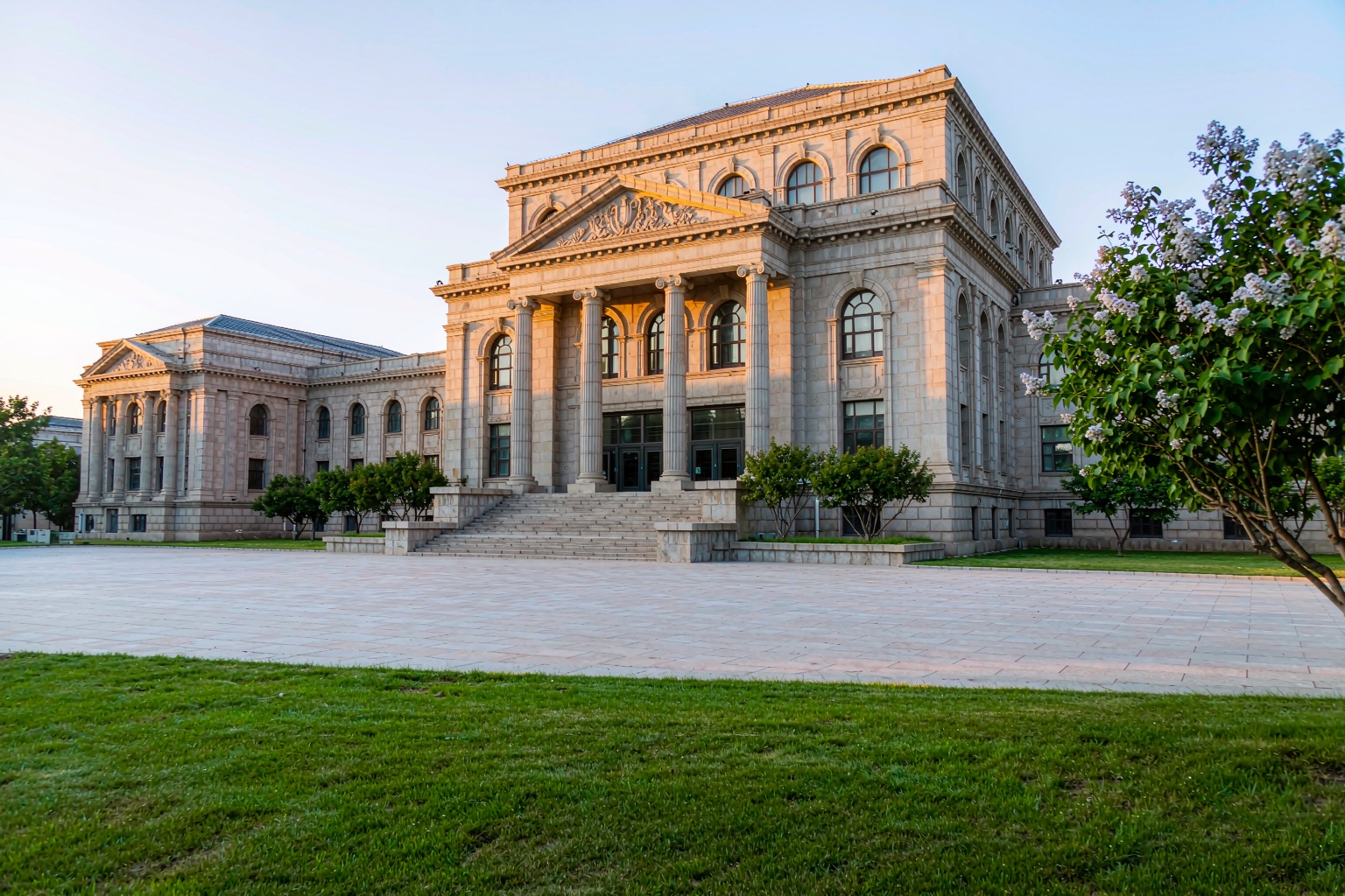 The neoclassical stone hall of the Lushun Museum behind a blossoming tree and mown lawn.