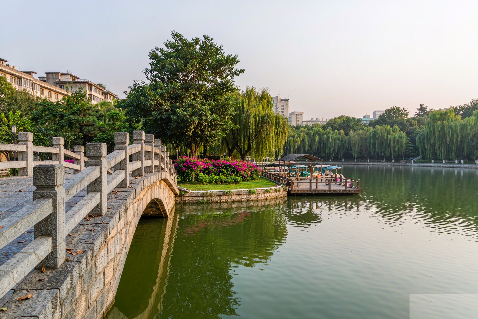 A stone arched bridge crossing the calm green water of Cuihu Park with willow trees and paddle boats, Kunming