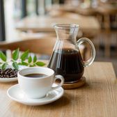 A glass carafe and white ceramic cup of black coffee on a light wooden cafe counter with a small dish of roasted beans