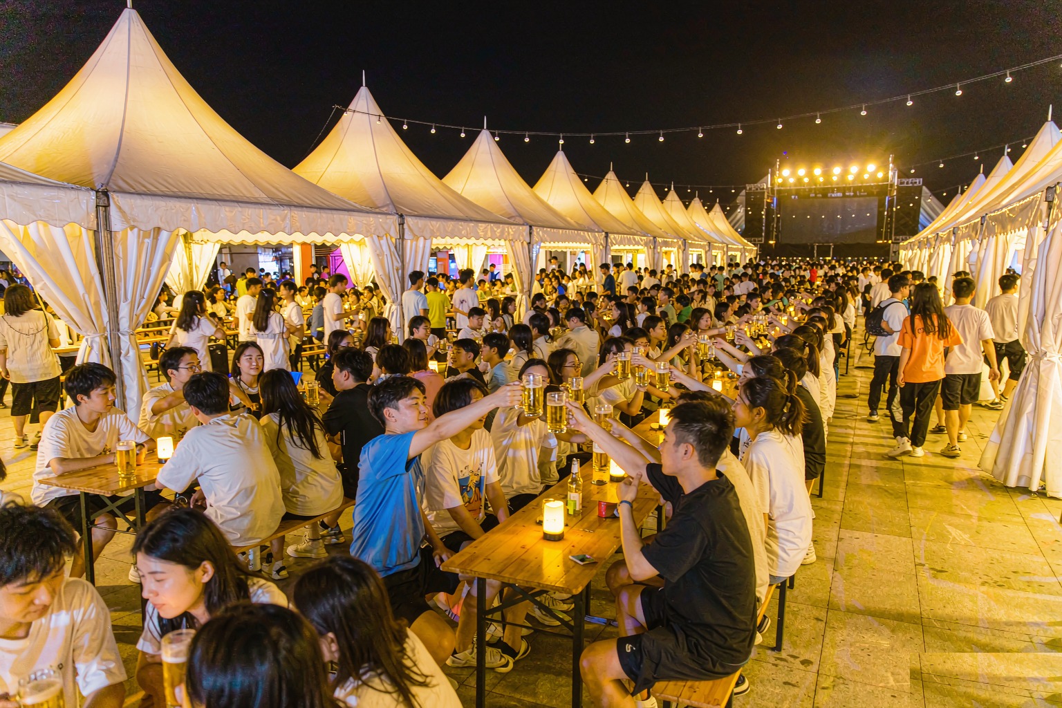 Rows of lit white marquee beer tents with shared wooden trestle tables and clinking mugs at the Qingdao International Beer Festival at night