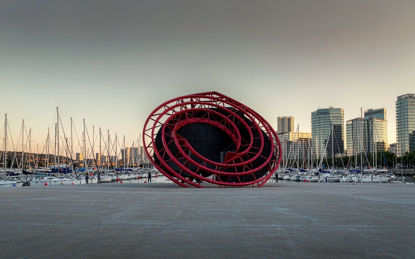 Red spiral sculpture on a waterfront with sailboat masts behind