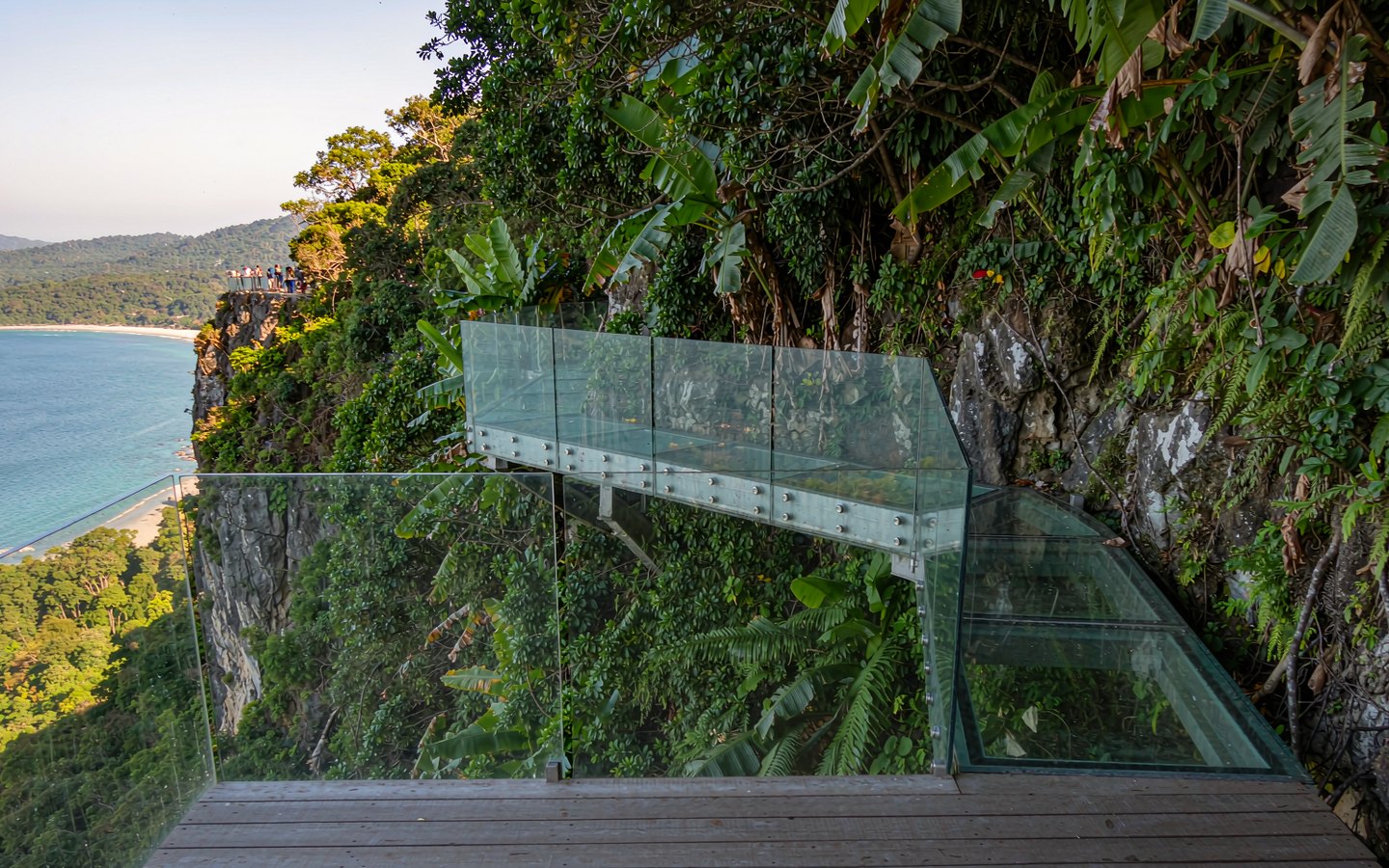 Glass walkway on a forested cliff above a wide turquoise bay