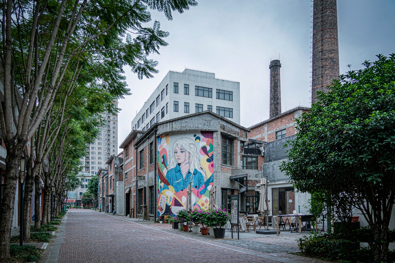 Red-brick former factory buildings with a colorful mural and chimneys along a tree-shaded lane at OCT-LOFT