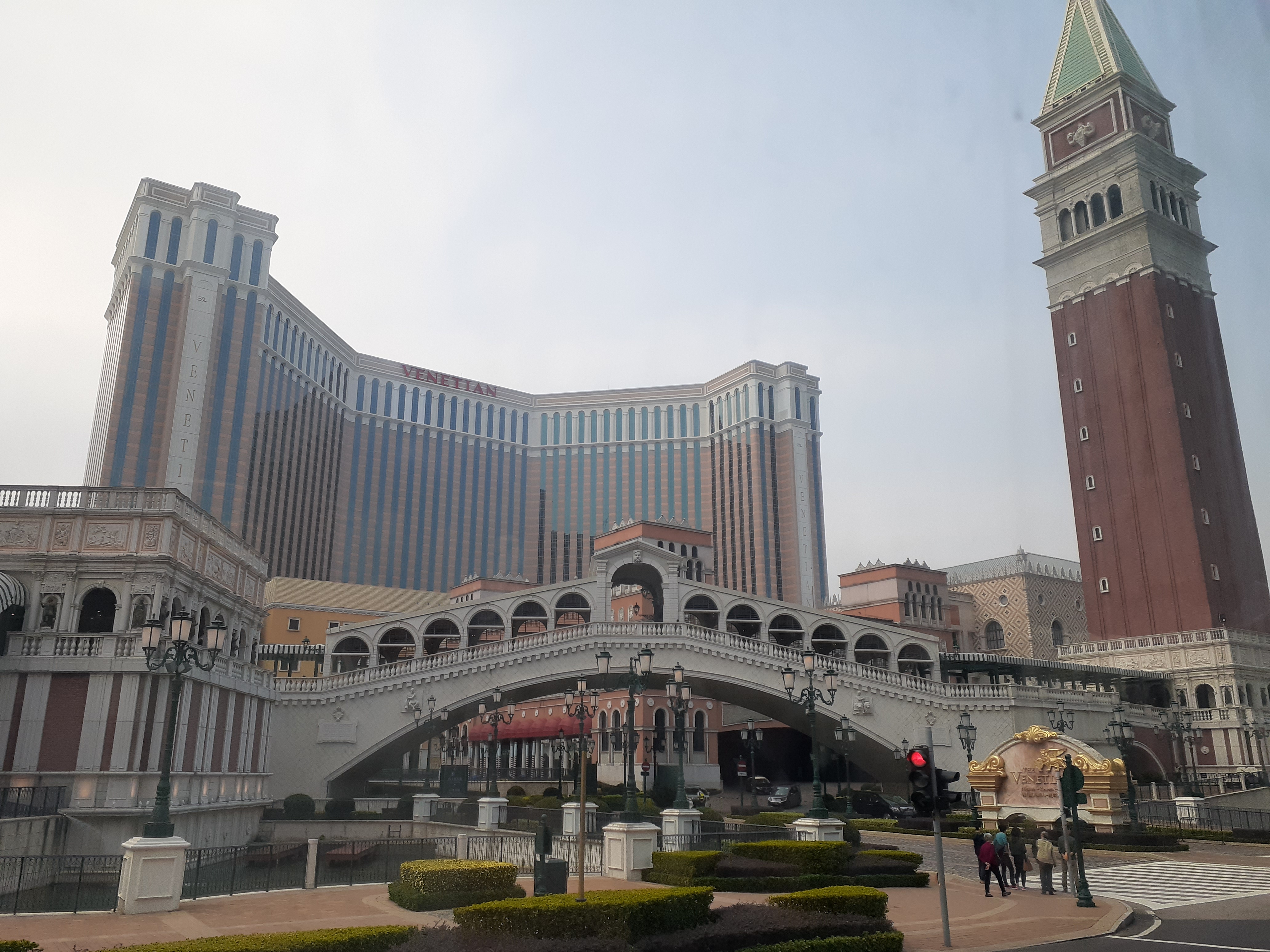 A replica Rialto bridge and brick campanile in front of the Venetian's striped hotel block, with clipped hedges and a crossing in front.