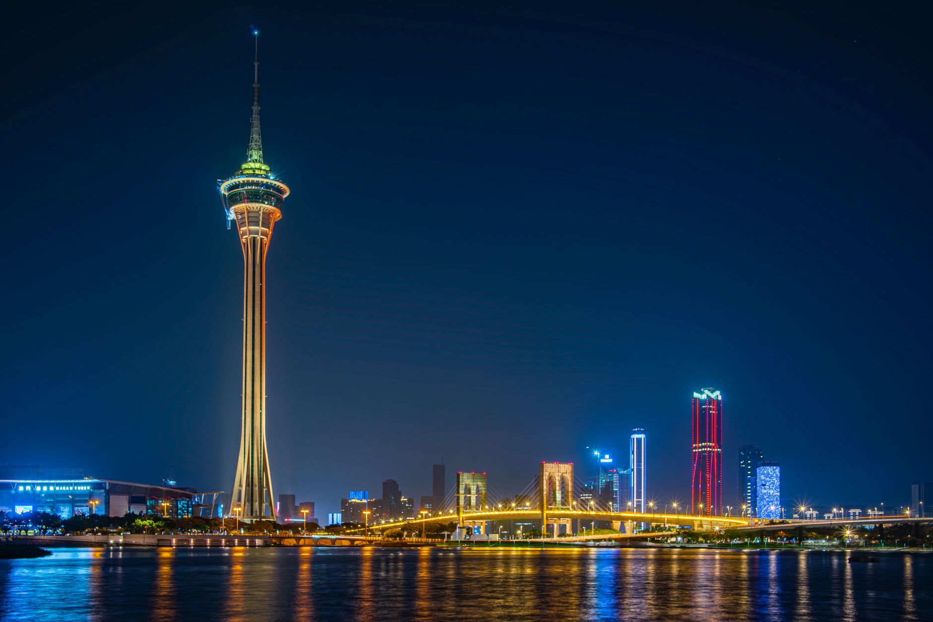 The lit Macau Tower and its observation ring above a cable-stayed bridge, with the Cotai skyline and its reflections behind.