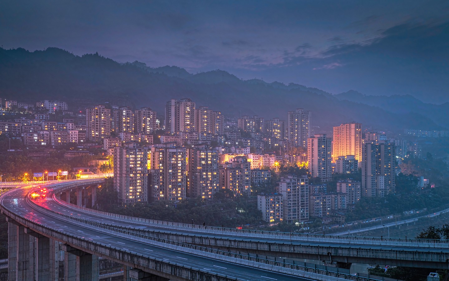 An elevated highway ramp curving past hillside apartment towers lit up at dusk in Chongqing