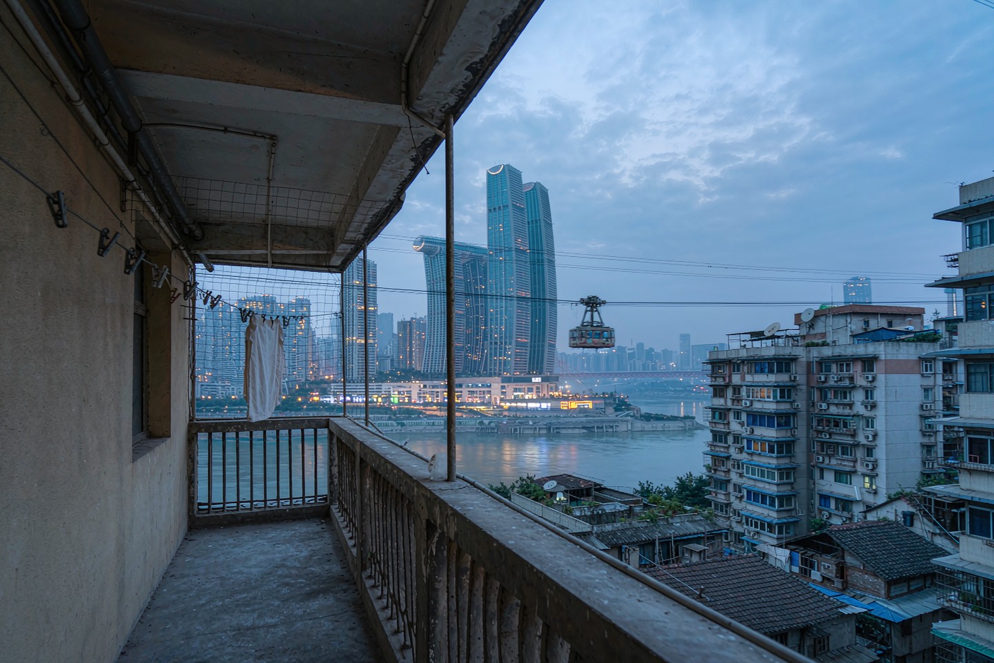 A concrete residential sky-bridge corridor with laundry lines, looking across a river to a glass tower skyline and a passing cable car at dusk